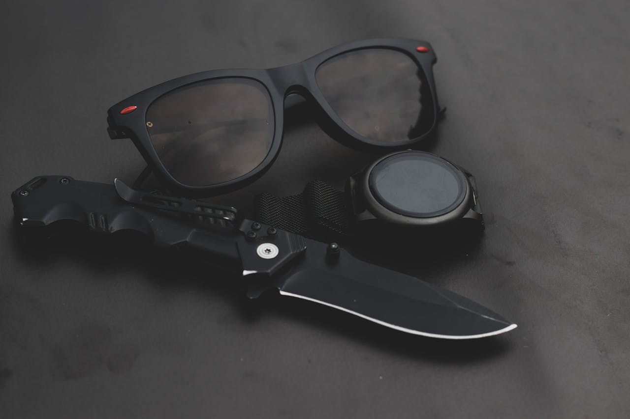 A minimalist black still life featuring sunglasses, a knife, and a wristwatch on a dark surface.